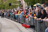 A crowd of people stand behind a gate, some waving Aboriginal and Torres Strait Islander flags