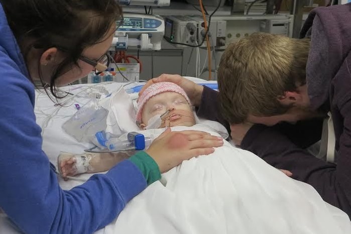 Two parents stand over a small baby on a hospital bed.