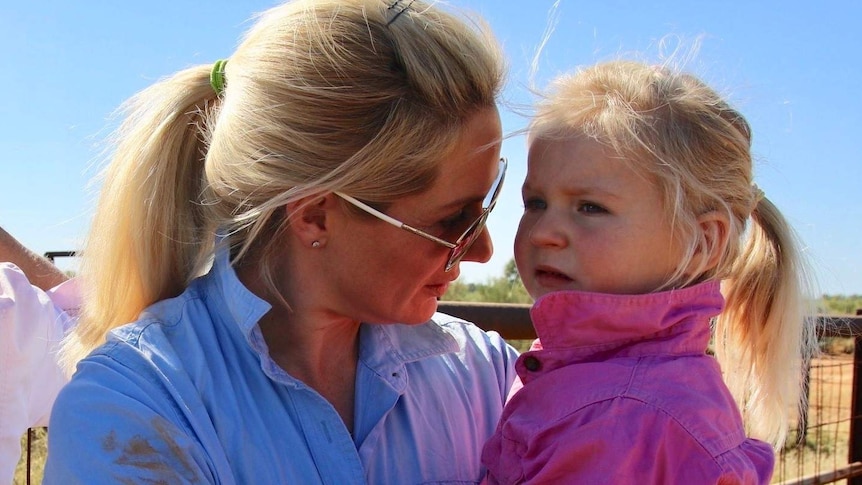 A woman with a blonde ponytail in a mud-smeared blue work shirt holds her young daughter who's wearing a pink work shirt.