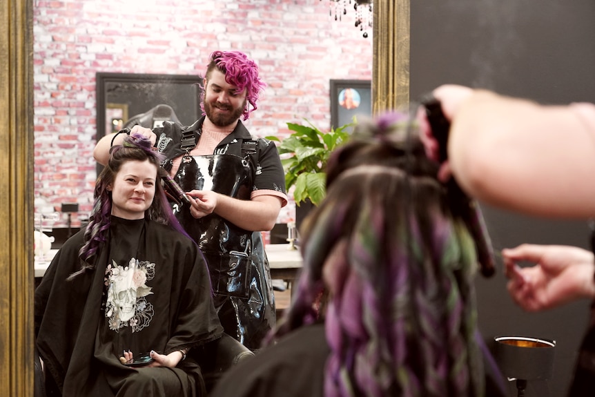 A woman with purple and green hair is at the hairdressers smiliing in the mirror.