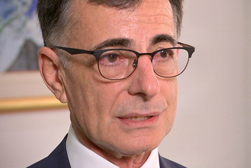 A headshot of a man wearing a suit and glasses, in front of a painting.