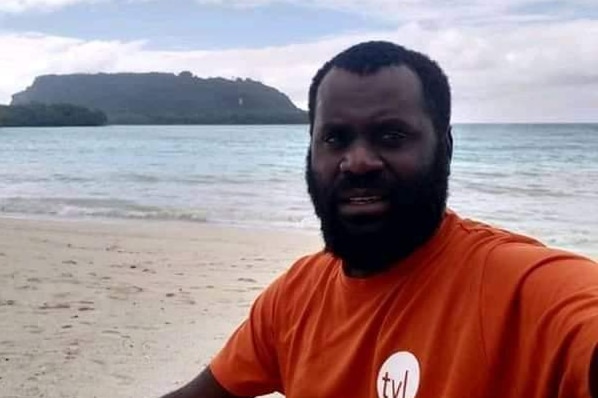 A ni-Vanuatu man in orange tshirt on a beach with island in the background