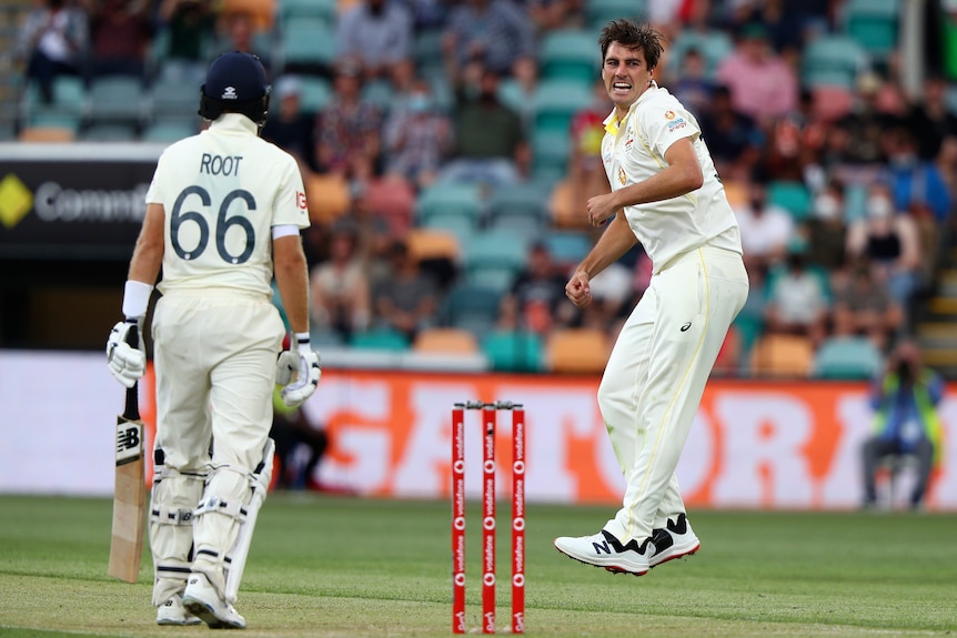 Australian bowler Pat Cummins shouts and jumps as England batter Joe Root stands in the foreground.