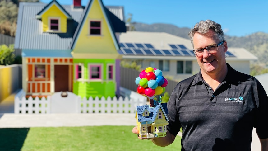 A man stands in front of a massive cubby house, holding a toy of the Pixar home that inspired it.