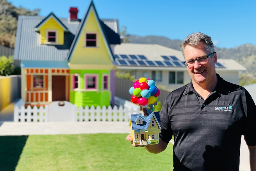 A man stands in front of a massive cubby house, holding a toy of the Pixar home that inspired it.