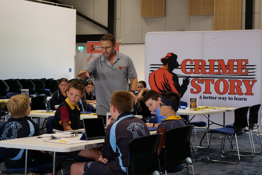 A teacher stands next to a group of students sitting at a desk with a large sign in the background saying "Crime Story".