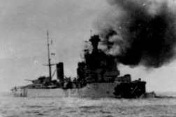 Historical photo of a stricken naval ship with smoke billowing out.