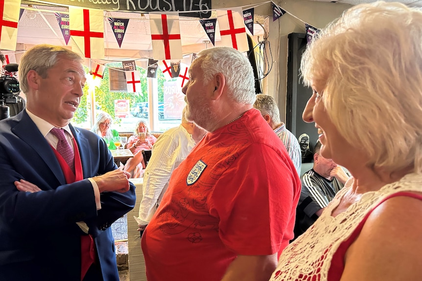 Nigel Farage stands in a suit speaking to a man and woman in a pub surrounded by English flags