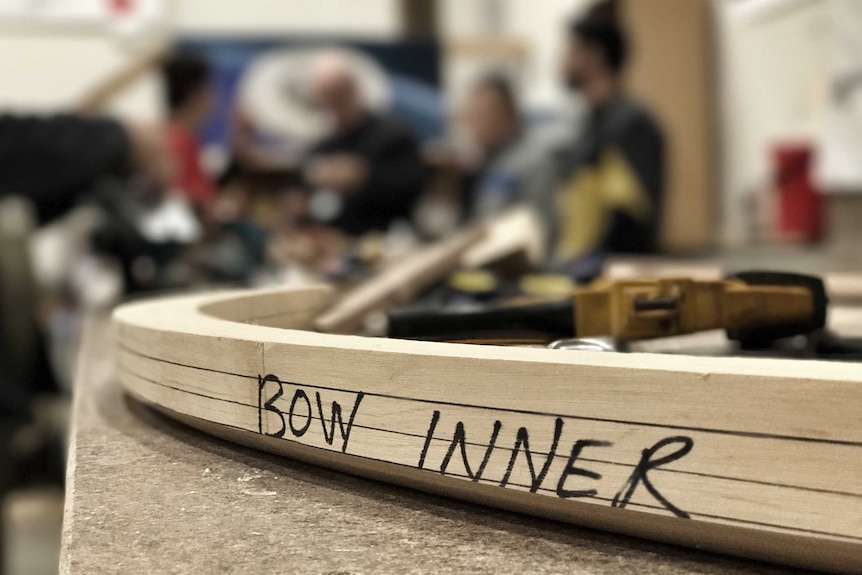 A close up of a boat piece called bow inner with refugee boat builders out of focus in the background in July 2017.