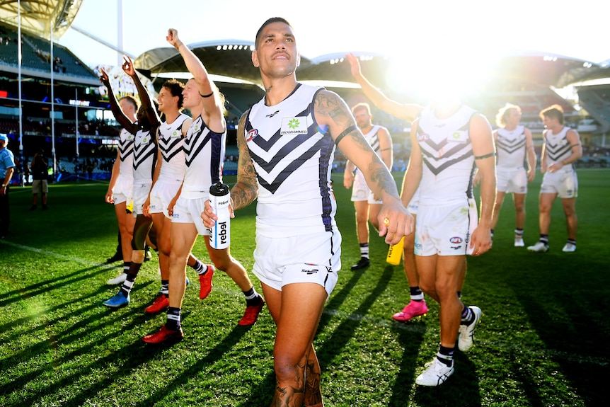 A Fremantle AFL player smiles and looks up at the grandstand as his teammates point in the air in celebration after a win. 