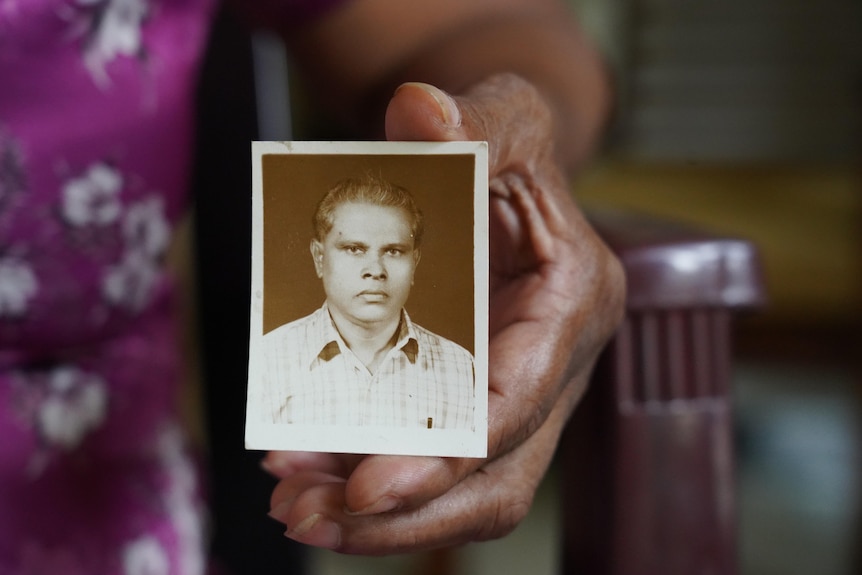 A woman holds a sepia-toned photograph up to the camera, of a young man with a stern expression