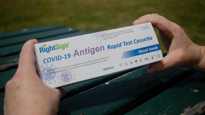 A person holds a thin white rectangular box with the words COVID-19 antigen test written on it.