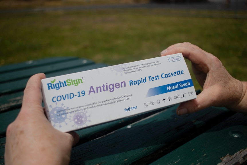 A person holds a thin white rectangular box with the words COVID-19 antigen test written on it.