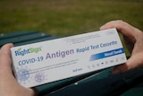 A person holds a thin white rectangular box with the words COVID-19 antigen test written on it.