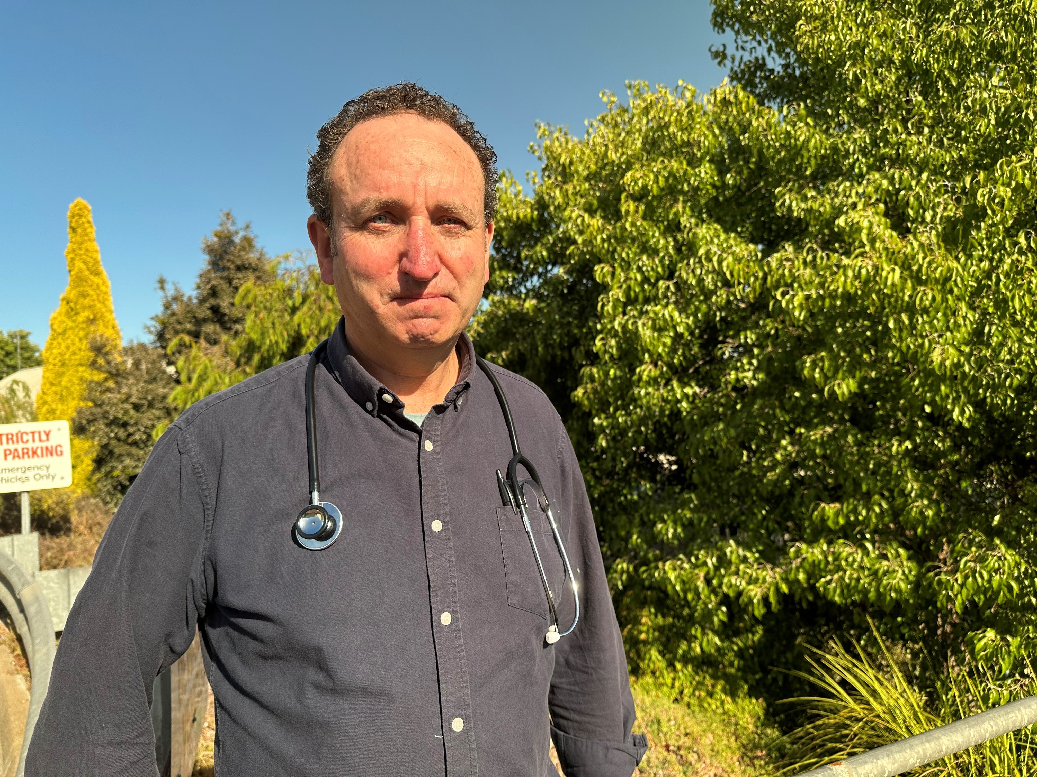 A male doctor stares into the camera with trees behind.