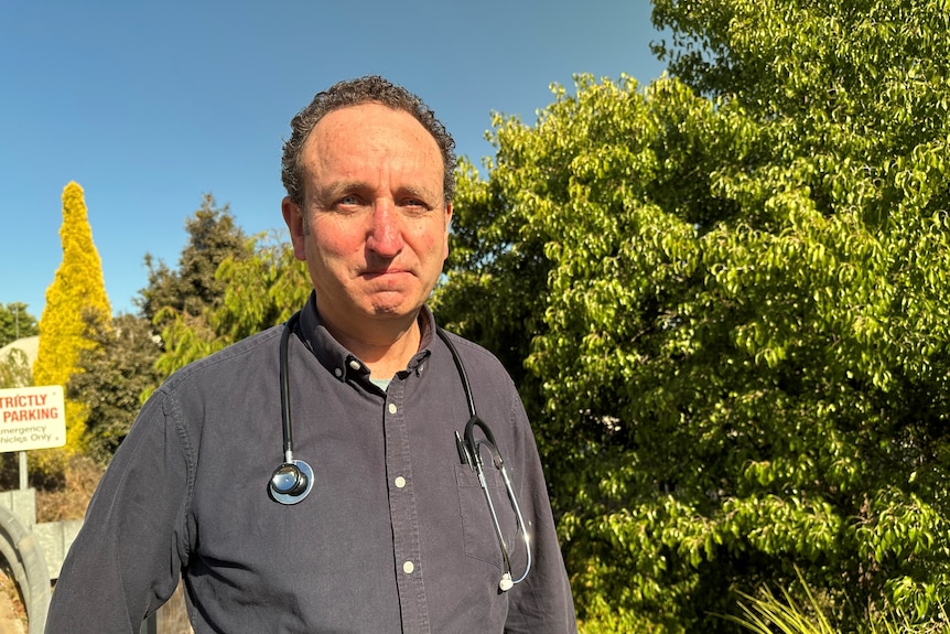 A male doctor stares into the camera with trees behind.