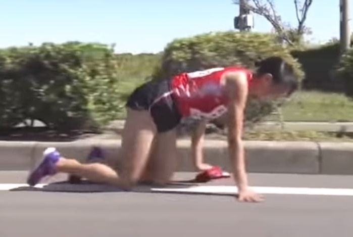 A woman dressed in running attire crawls on the a road on her hands and knees.