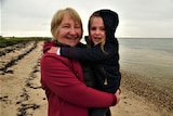 A grandmother with short blonde hair wearing a red coat stands on a beach and holds her 4yo granddaughter on her hip.