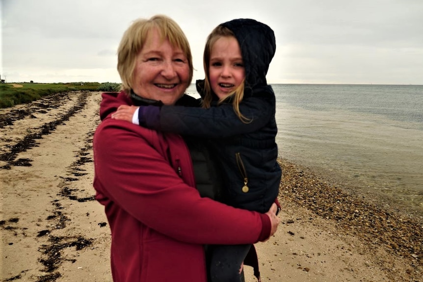 A grandmother with short blonde hair wearing a red coat stands on a beach and holds her 4yo granddaughter on her hip.