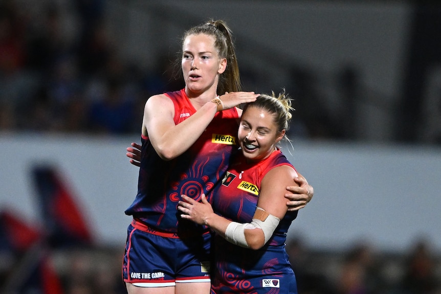 A tall young woman in a red and blue AFLW uniform hugs a less tall young woman in the same AFLW uniform on a football field.