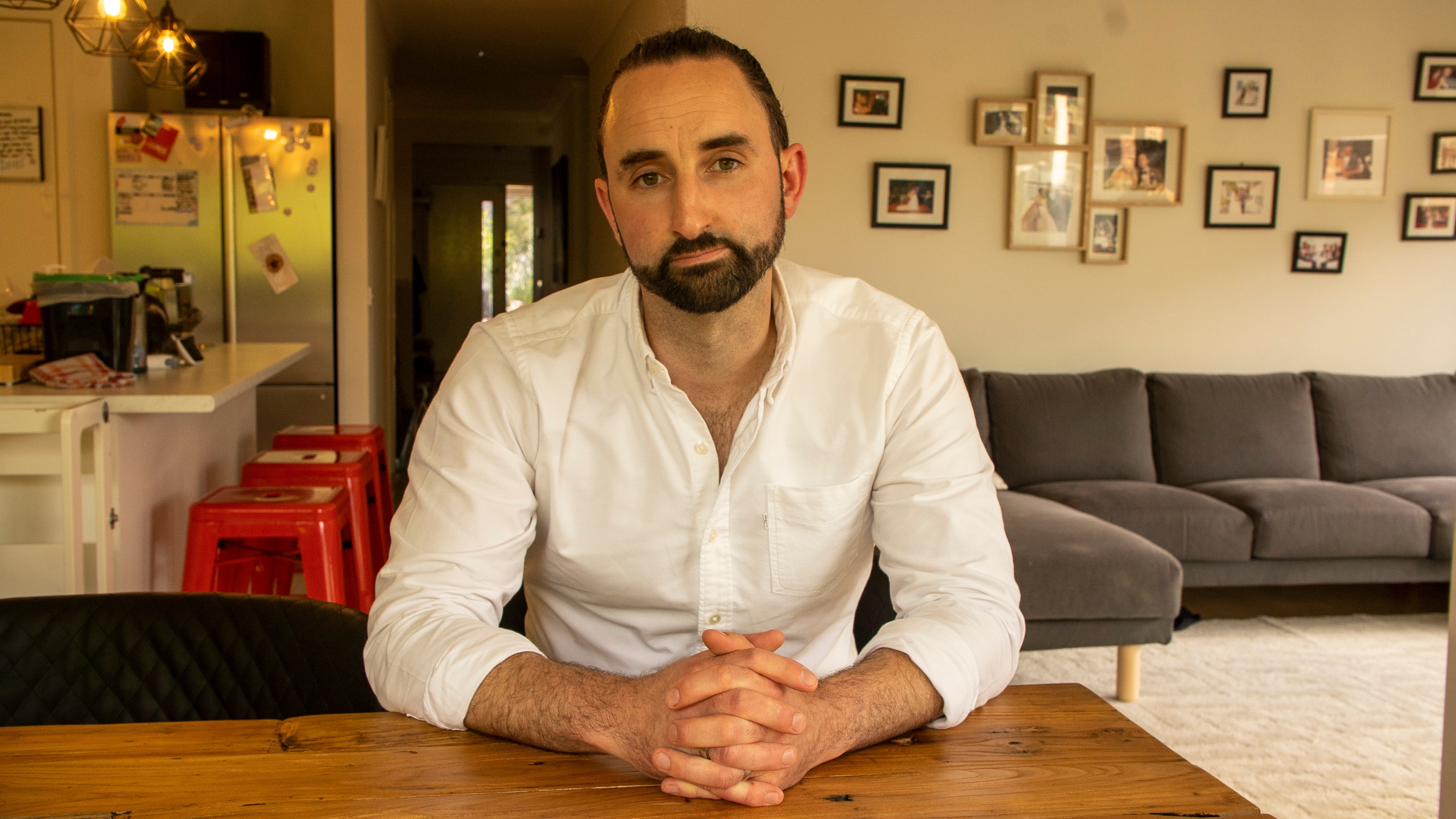 A young man with short dark hair and a beard sits at his dining table looking into camera with a serious expression.