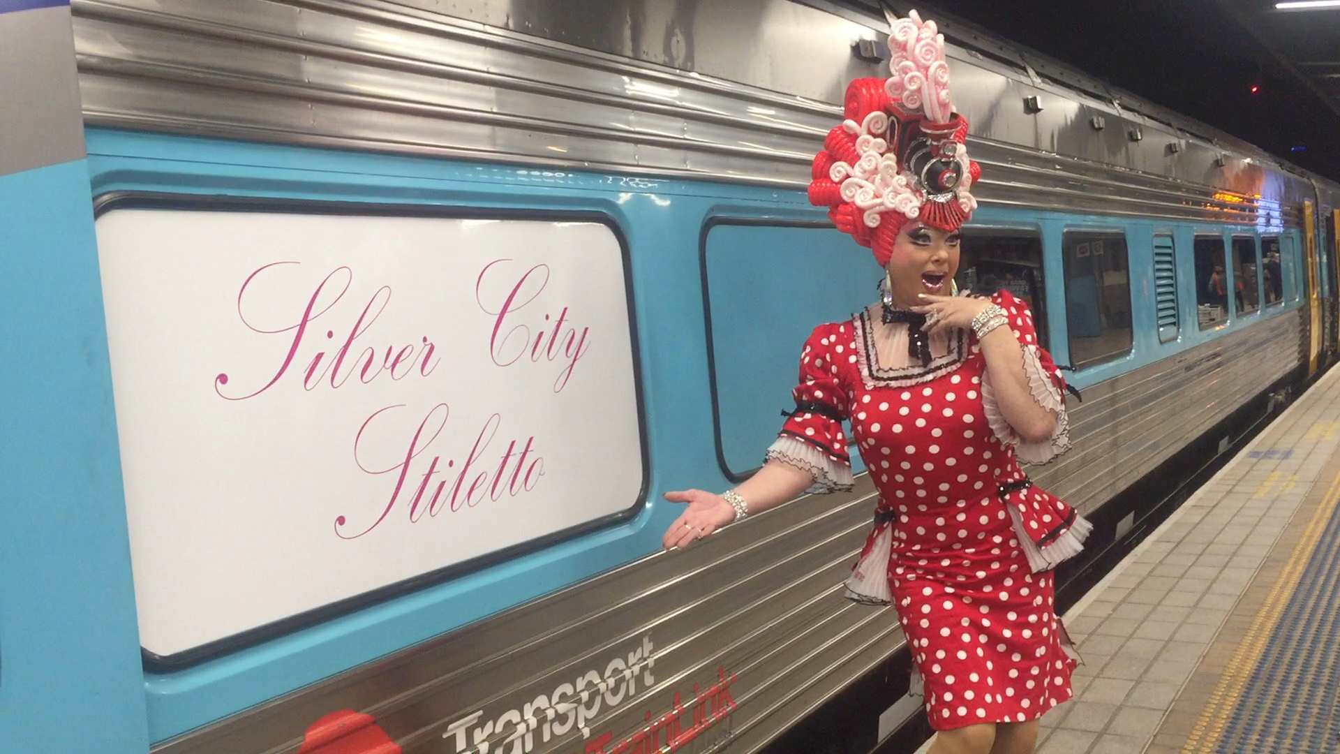 A drag queen poses with a sign on a train reading &#x27;silver city stiletto&#x27;.