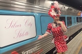 A drag queen poses with a sign on a train reading 'silver city stiletto'.