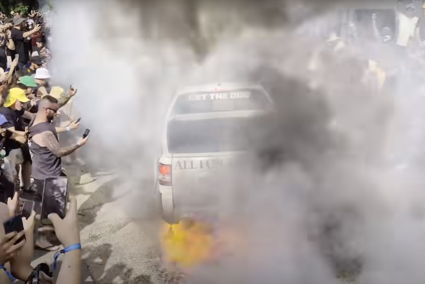A ute doing a burnout surrounded by a crowd of people. 