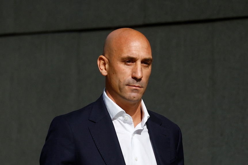 Luis Rubiales walks past a grey background with a serious expression 