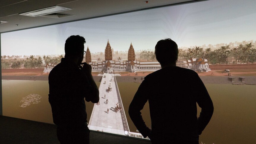 Two men stand silhouetted in front of a projection of virtual Angkor Wat