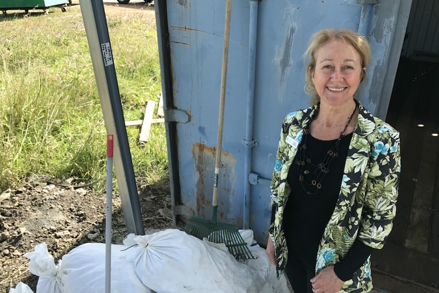 A woman with blonde hair, wearing a floral jacket, stands beside white plastic bags filled with oyster shells.