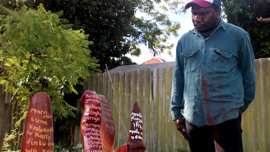 A young Aboriginal man in a baseball cap, denim shirt and black pants looks at wooden artworks with words inscribed on them.