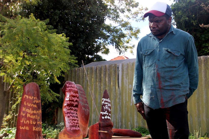 A young Aboriginal man in a baseball cap, denim shirt and black pants looks at wooden artworks with words inscribed on them.