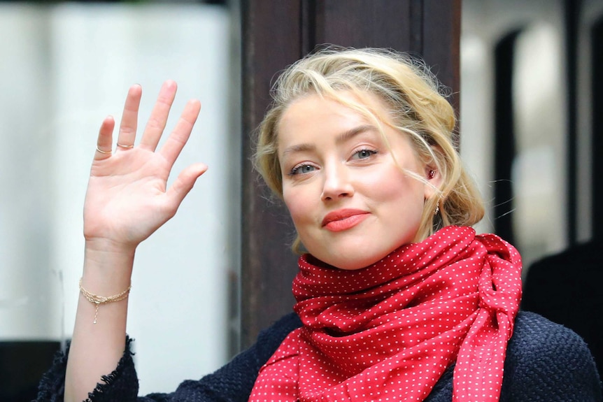 A blond woman waves, she is wearing a red scarf around her neck.