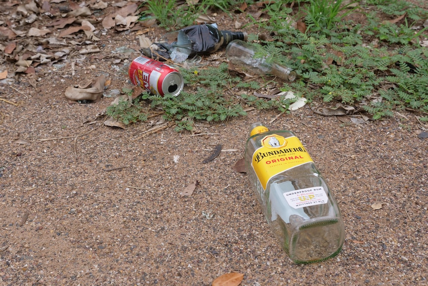Une canette de bière vide, une bouteille de spiritueux et une bouteille de vin cassée sur le sol.