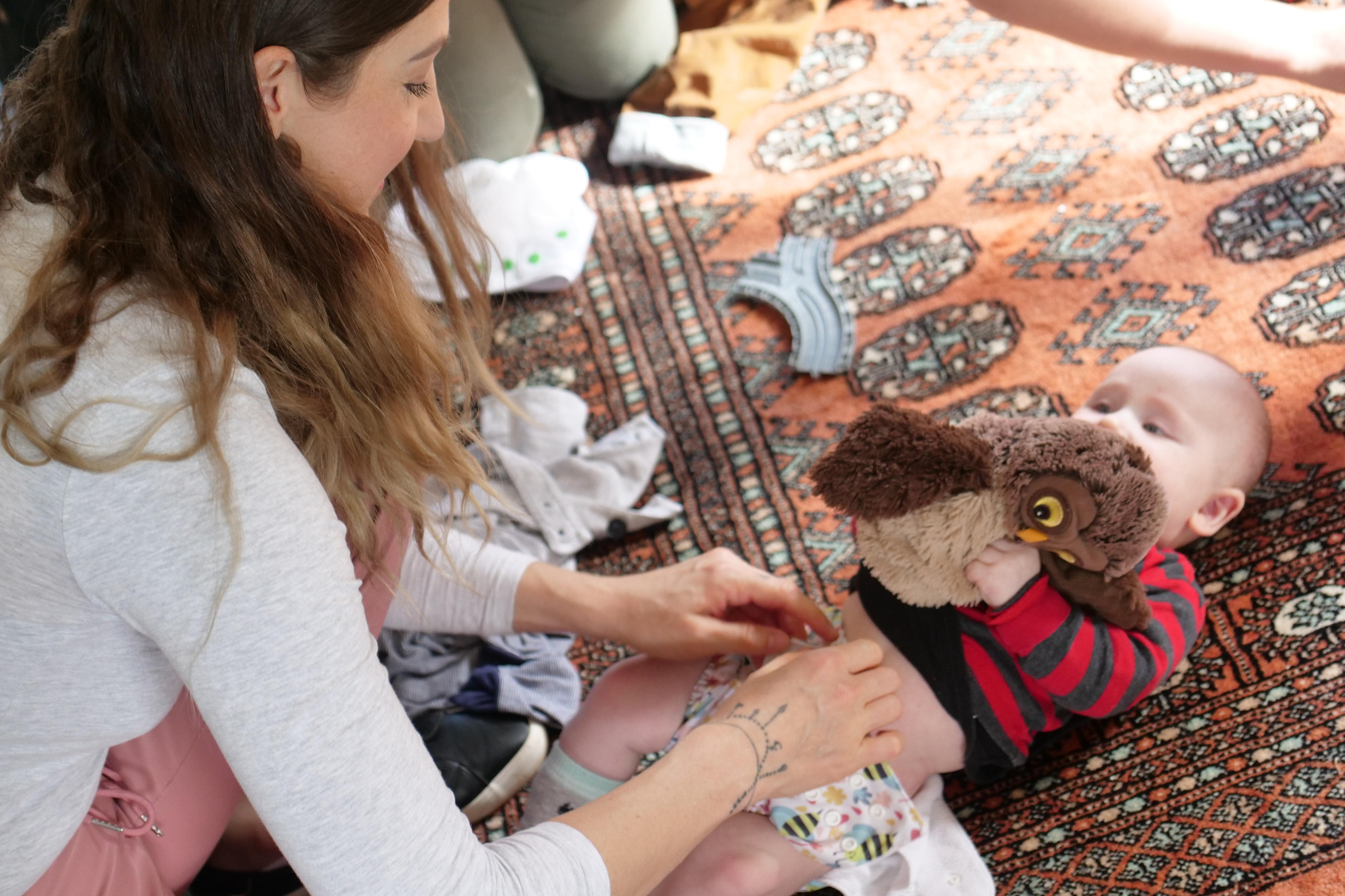 Mother changes her baby's nappy while he holds an owl teddy.