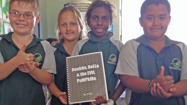 Four children in school uniforms hold a book and smile for the camera.