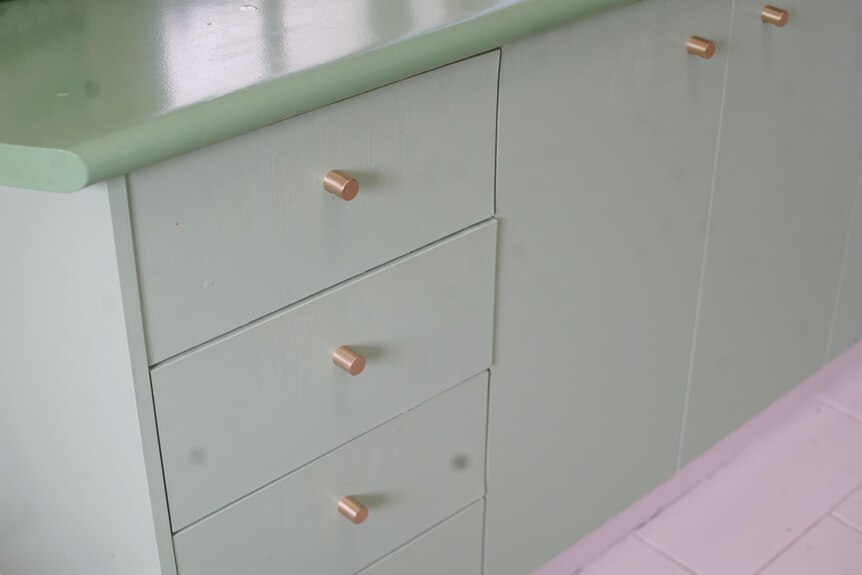 A pea green kitchen countertop is seen with mint green kitchen cupboards and drawers with gold handles.