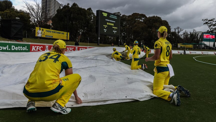 Australia's women's cricket team crouch trying to grab the covers with dark clouds overhead as play is stopped for rain.