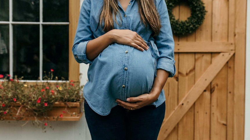 A woman in a denim shirt holding her pregnant belly.