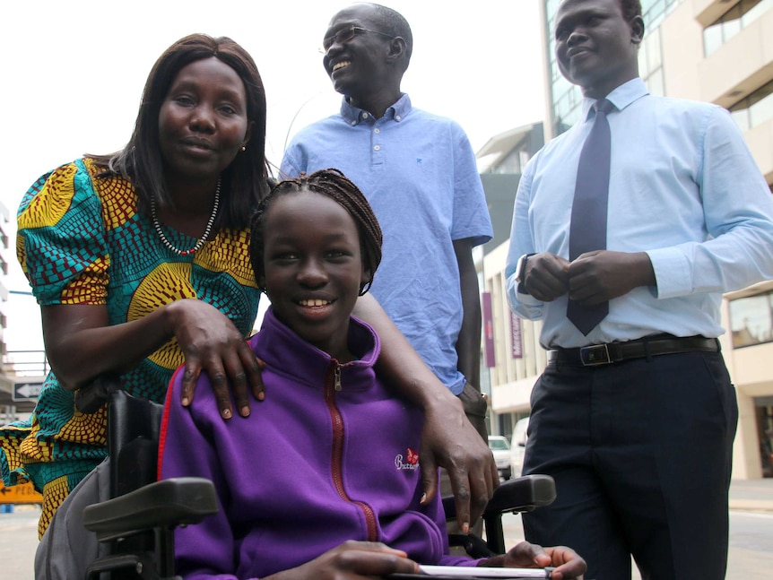 Sunday sits in a wheelchair smilling, with her mother behind her, and two men to the right, also smiling.