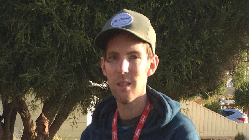 A thin man in his early 30s wears a blue hoodie and baseball cap.