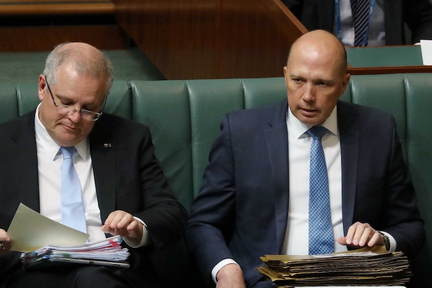 Two white men wearing blue ties and suits sit on a green bench.