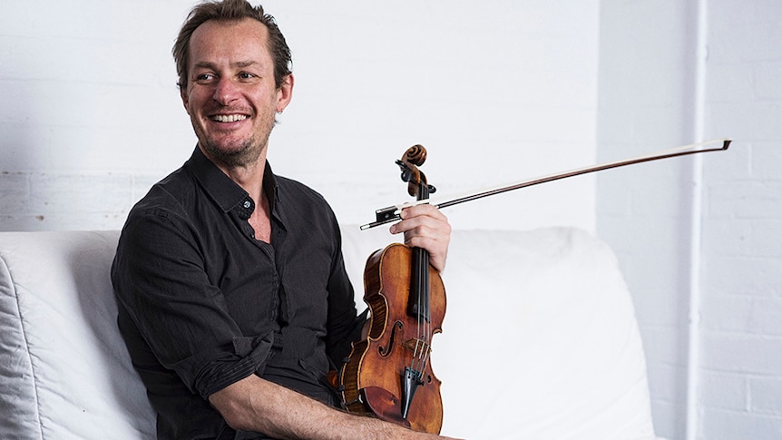Richard Tognetti sits on a while lounge holding his violin. He looks to one side smiling.