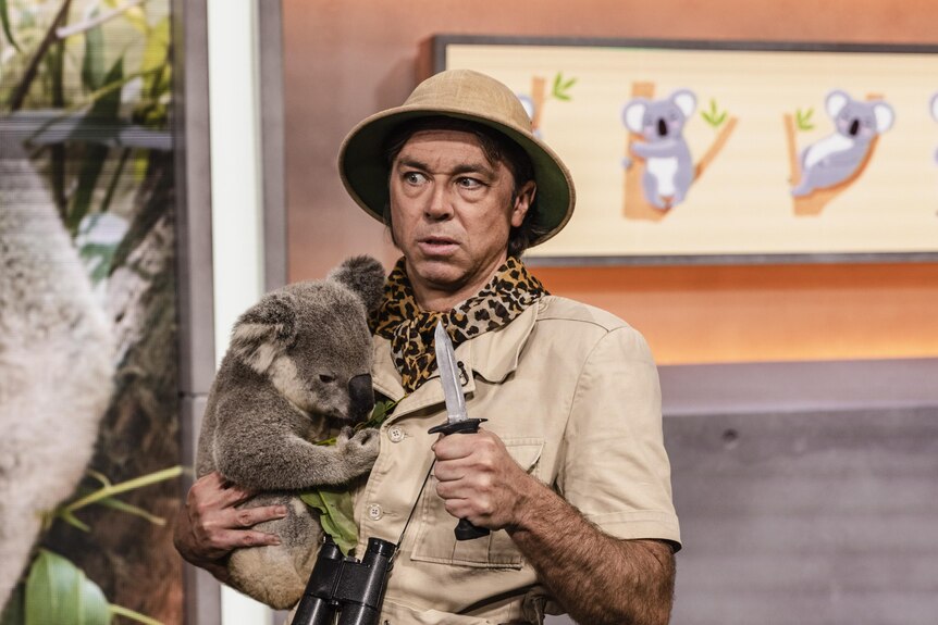 Kick Gurry in Caught holding a knife in one hand and a koala in the other. He's wearing a hat and wildlife uniform