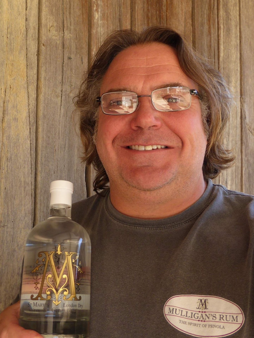 A man with glasses and a grey t-shirt reading "Mulligan's rum the spirit of Penola" holds a bottle filled with clear liquid