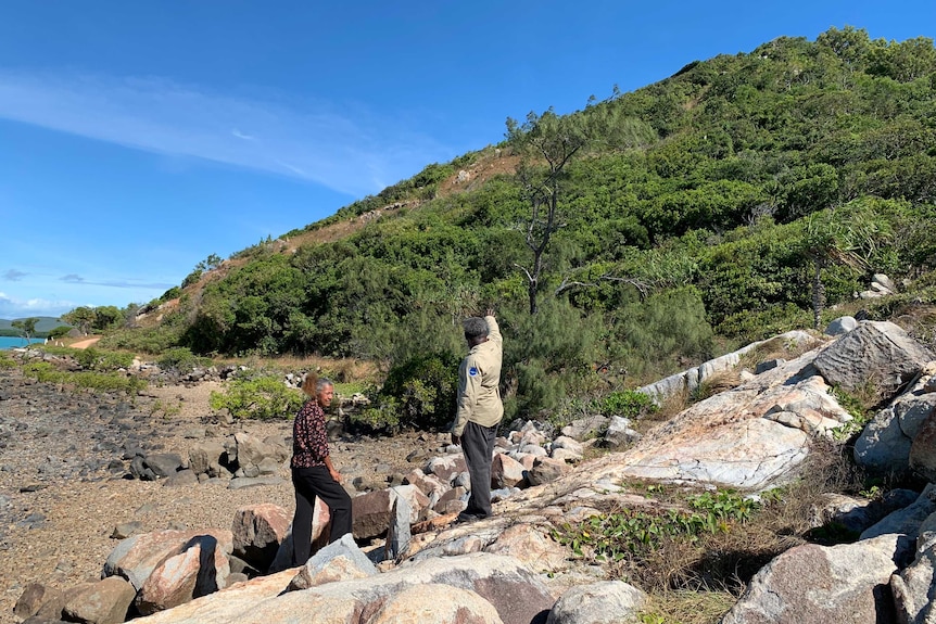 Enid Tom and Milton Savage stand on rocks at the foot of a hillside. Milton is pointing at the hill.