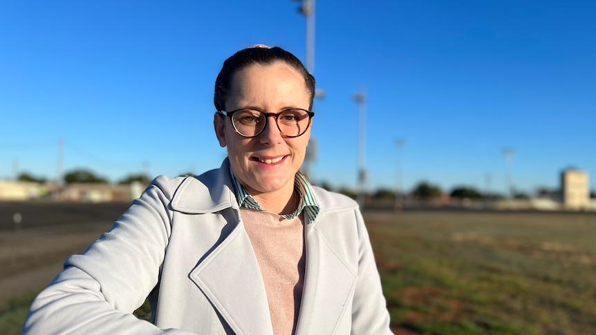 A woman wears a pale jacket with a field behind her.