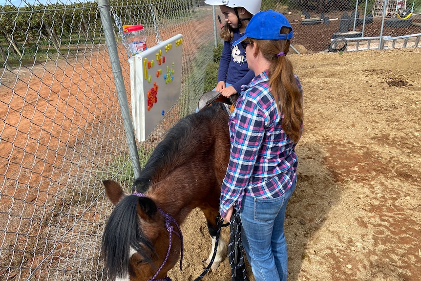 A girl sits on a horse with a woman beside her at a fence with a letter board.