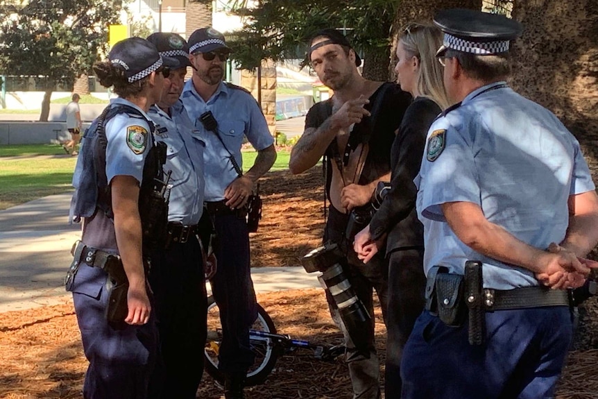 A man with a camera talks to four police who stand around him in a circle